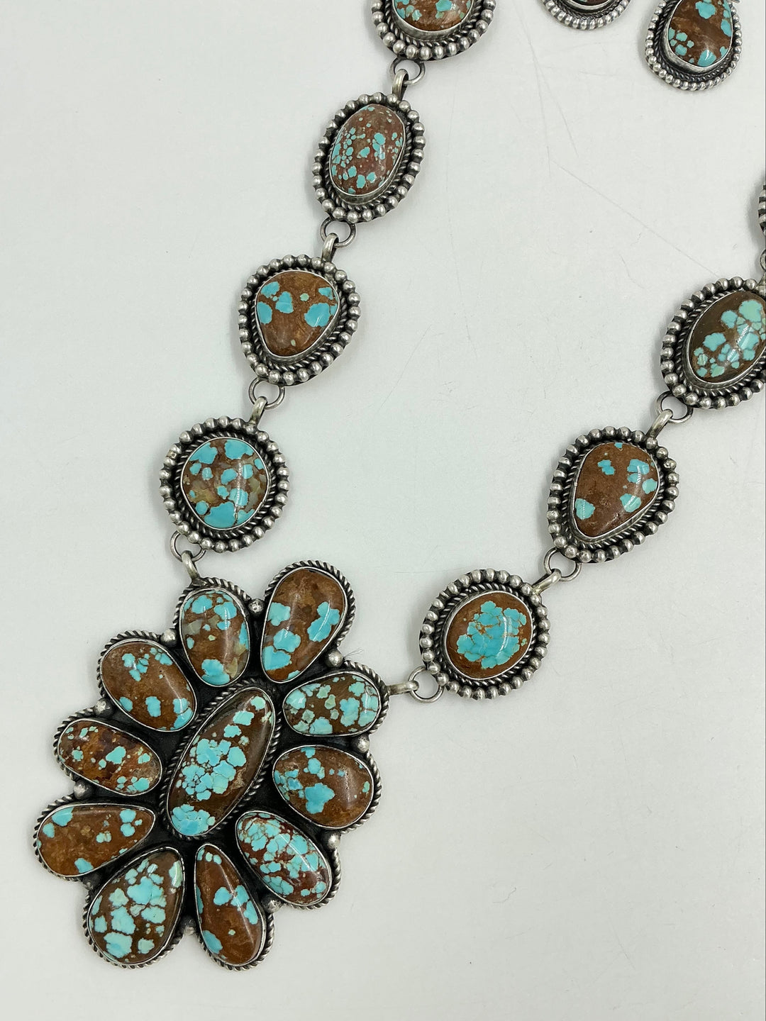Turquoise Necklace with brown and blue stones in a center cluster of many in a flower and side stones set in sterling silver on a chain with a white background and earrings. 