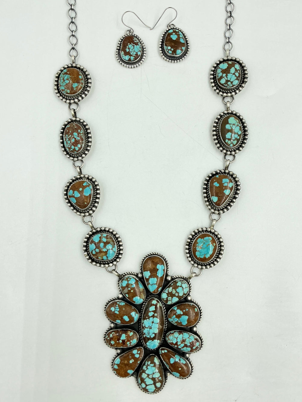 Turquoise Necklace with brown and blue stones in a center cluster of many in a flower and side stones set in sterling silver on a chain with a white background and earrings. 