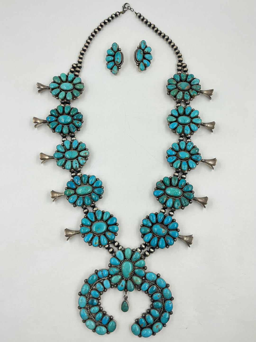 Kingman turquoise squash blossom necklace and earrings set with teal colored stones on a white background