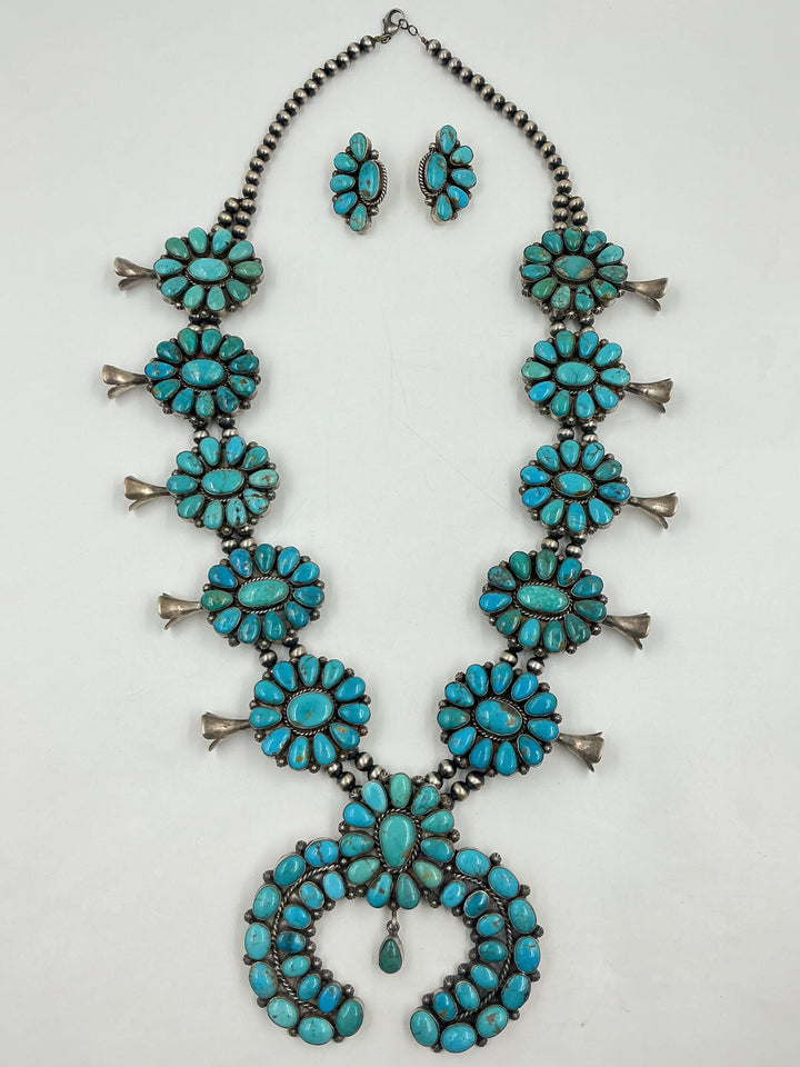 Kingman turquoise squash blossom necklace and earrings set with teal colored stones on a white background