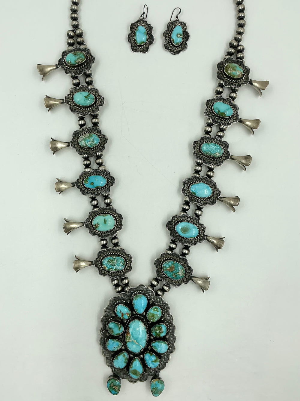 Squash blossom necklace and earrings set with blue/green stones on a white background