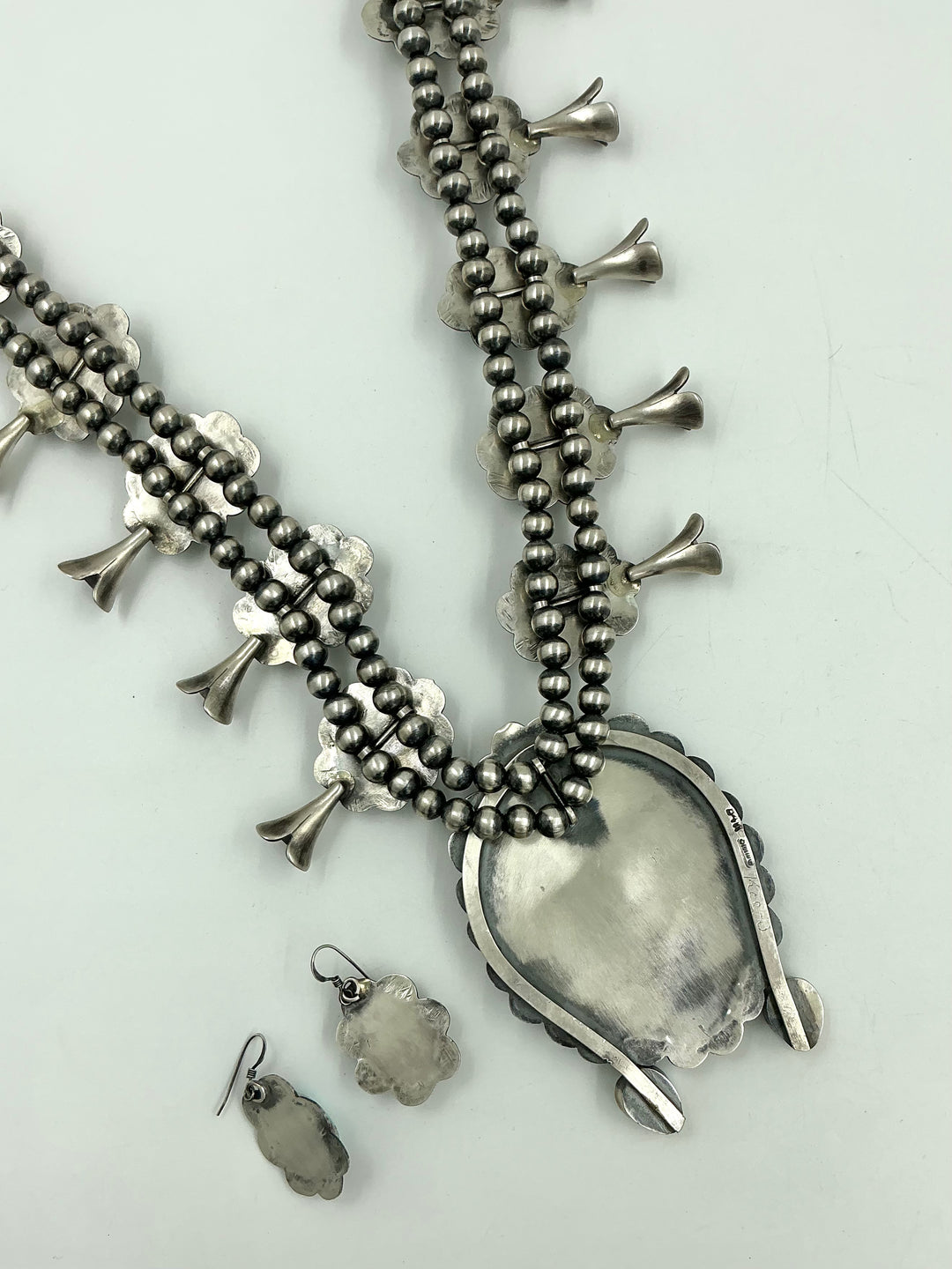 Silver back of squash blossom necklace and earrings set with blue/green stones on a white background